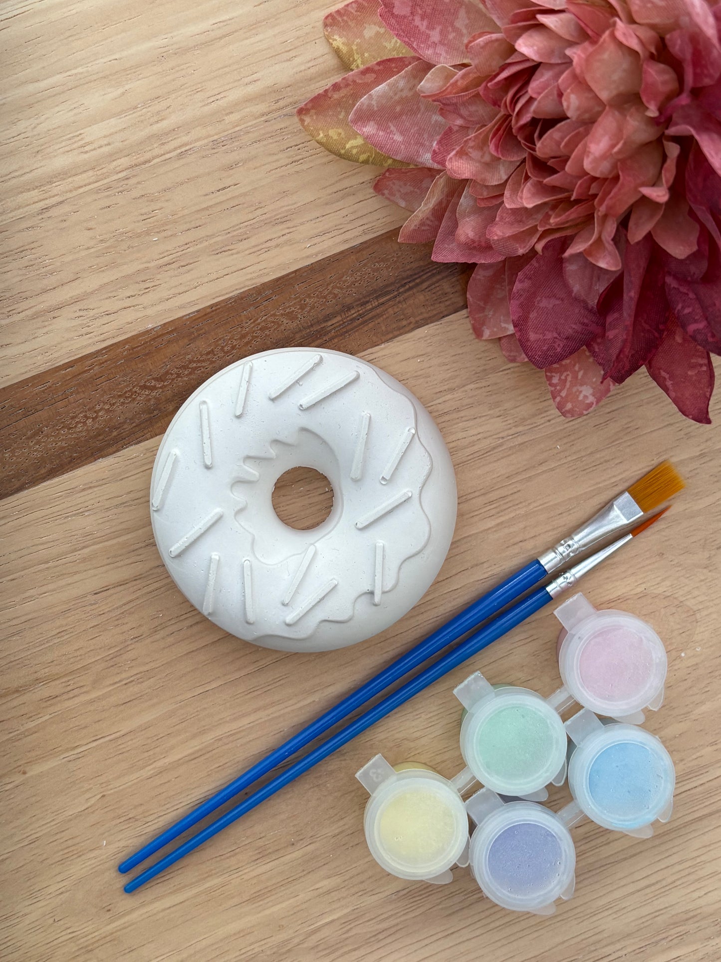 A white ceramic donut painting kit shown on a light wood background, featuring a painted donut base, two blue-handled paintbrushes, and five small paint pots in pastel colors. A pink flower is visible in the top right corner.