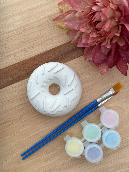 A white ceramic donut painting kit shown on a light wood background, featuring a painted donut base, two blue-handled paintbrushes, and five small paint pots in pastel colors. A pink flower is visible in the top right corner.